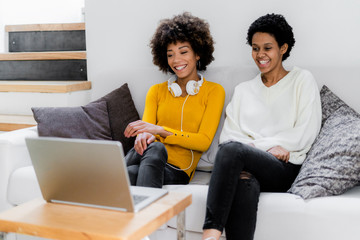 Portrait of two laughing friends sitting side by side on the couch watching movie on laptop