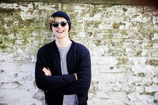 Portrait of laughing young man with headphones wearing sunglasses and cap in front of weathered wall