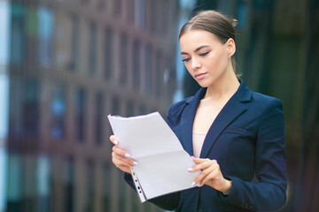 Beautiful confident business woman, young European gorgeous girl in jacket standing outdoors, reading, looking at documents, papers or contract with serious look. Attractive office worker, lady.