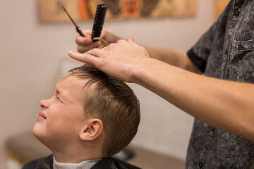 A pretty boy toddler happy to be on the haircut with a professional children's hairdresser. Blond little boy having a haircut at hair salon. Hairdresser's hands making hairstyle to child.