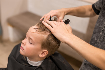 A pretty boy toddler happy to be on the haircut with a professional children's hairdresser. Blond little boy having a haircut at hair salon. Hairdresser's hands making hairstyle to child.