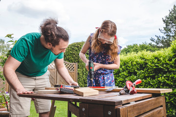 Young man looking at sister drilling on plank while standing in yard