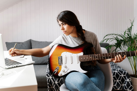 Young woman writing while using laptop and electric guitar at home
