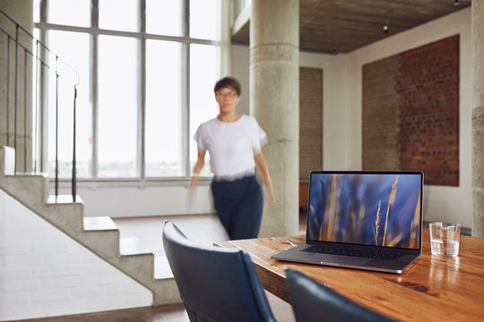 Laptop on wooden table in a loft flat with woman walking in background
