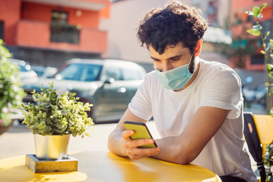 Young man wearing face mask using smart phone while sitting at sidewalk cafe