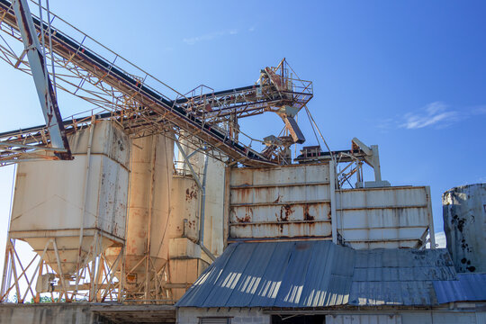 Horizontal view of conveyors, silos, hopper, and tin roofing of an old concrete plant against the background of a blue sky..