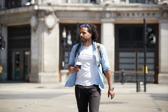 Portrait of young man with coffee to go crossing the street while listening music with earphones, London, UK