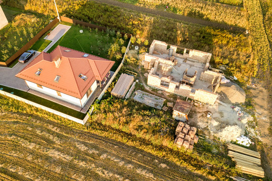 Top down aerial view of two private houses, one under construction with concrete foundament and brick walls and another finished with red tiled roof.