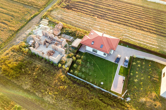 Top down aerial view of two private houses, one under construction with concrete foundament and brick walls and another finished with red tiled roof.