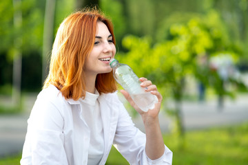 Happy thirsty redhead woman drinking fresh bottled water in summer outdoors.