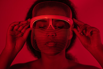 Close-up of young woman wearing red led mask against wall at home