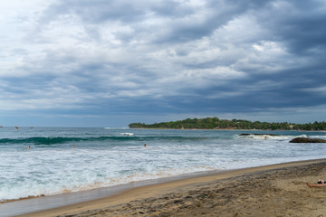 Tropical beach with waves, palm trees  and rocks in the sea. The sky is cloudy. Arugam bay, Sri Lanka