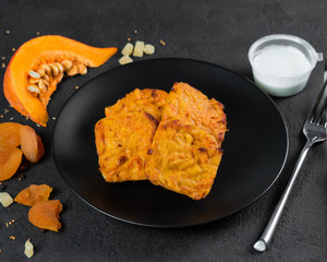 Vegetarian breakfast. Pancakes of pumpkin, dried apricots and dried fruits are on a black plate. Next to it is a piece of pumpkin and dried apricots. White cream sauce. A black stone background. Pumpk