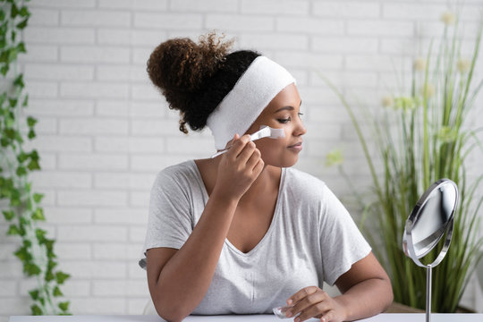 Young Woman Wearing Headband Applying Facial Mask While Looking In Mirror At Home