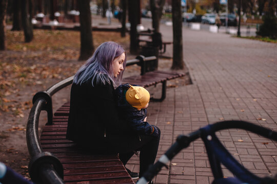 Young mother holding baby son while sitting on bench in park in autumn