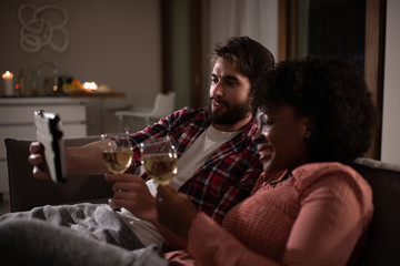 Multiracial man and woman with glasses of wine smiling and making video call to friends while resting on couch during romantic date at home