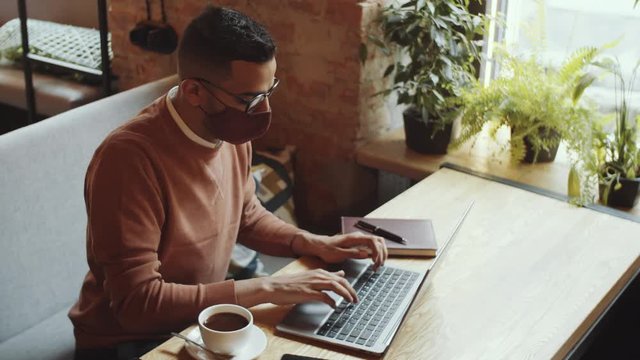 High angle shot of young middle eastern man in face mask sitting at cafe table with coffee cup on it and using laptop while working remotely during covid-19 pandemic