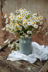 Bouquet of wild chamomiles in old metal can on wooden background with old scissors, white towel and orange flowers. Country still life.