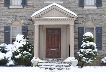 Elegant wooden front door in portico entrance of house with snow covered bushes