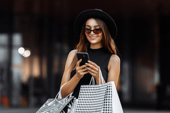 Happy beautiful elegant girl in a black dress and hat, using a mobile phone, holding shopping bags. Black Friday, shopping