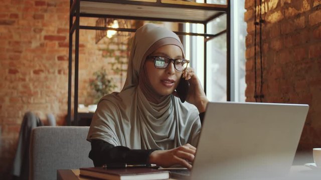 Beautiful muslim businesswoman in glasses and hijab talking on mobile phone while sitting at cafe table with laptop on it