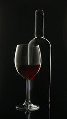 glass goblet with red wine next to a black glass wine bottle on a black background with contour lighting