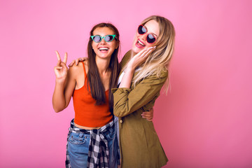Lifestyle cheerful portrait or two stylish pretty woman posing together, hugs, showing v science and sending air kiss, trendy 90s style outfit, cute emotions, friendship concept.