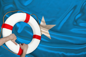 male hands hold on to a white life buoy against the backdrop of the silk national flag of the country of Somalia, the concept of medical insurance, tourism, disaster, humanitarian assistance