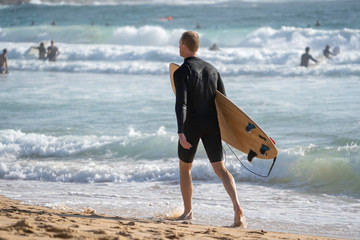 Surfer walking along the beach with his surfboard under his arm, looking for a place to go for a relaxing surf