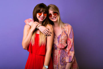 Friendship and communication concept, two sisters best fronds girls wearing colocar matching trendy summer dresser hugs and looks on camera, boho glasses, purple background.