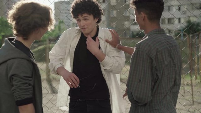Scared Caucasian teenager bullied by confident multiethnic friends at mesh fence. Portrait of shy frightened young man talking to multiracial hooligans in urban district in 1980s or 1990s.