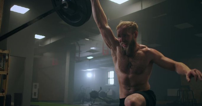 The boy pushes the barbell in the gym with one hand. An athlete without a T-shirt in a dark room lifts the barbell while practicing the force of the impact