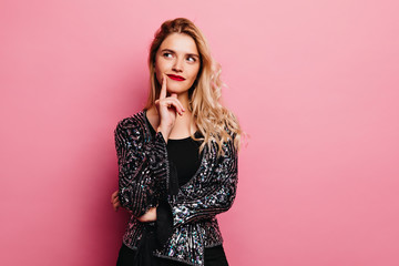 Adorable blonde girl posing with pensive face expression. Pretty young woman in sparkle jacket standing on pink background.