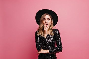 Surprised glamorous woman looking to camera. Enthusiastic pretty girl in black hat standing on pink background.