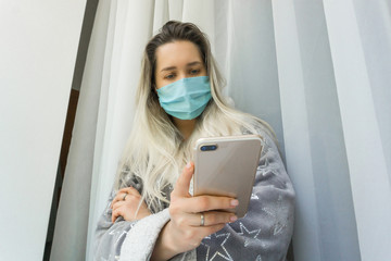 Woman wearing protective mask and holding, reading and checking online news on her smart phone at home office. Looking at telephone screen and worried, sad face. Global pandemic and self quarantine.