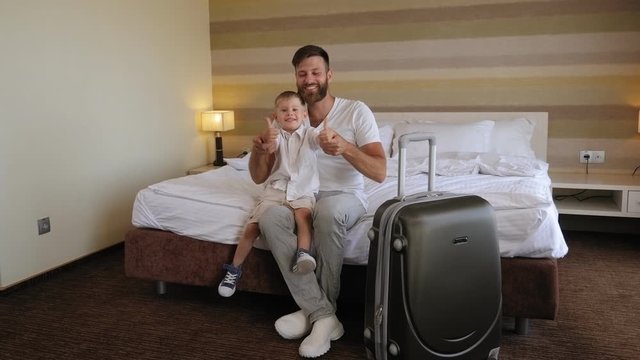 A happy bearded man sits with his little son on a hotel bed next to a large gray suitcase, they give a thumbs-up.