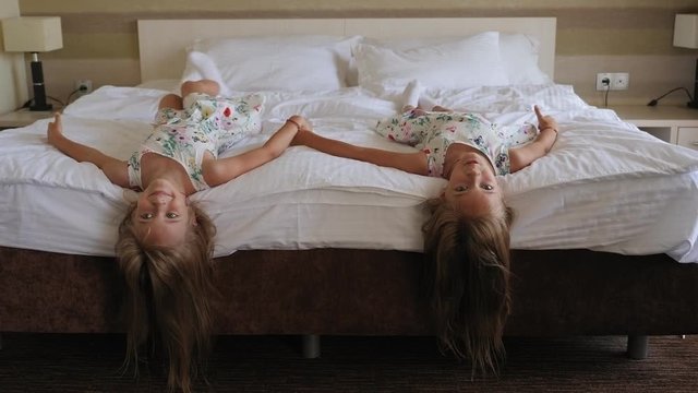 Two twin girls in identical dresses are lying on the bed upside down in a hotel bedroom and laughing.