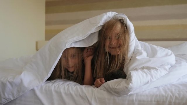 Two funny twin girls are playing at home on the bed, they are covered with a blanket and look out from under the blanket.