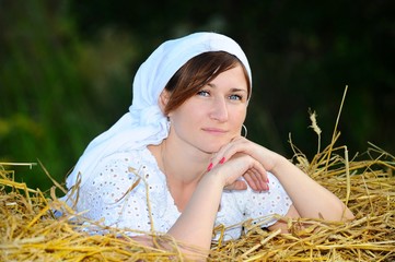 A peasant girl in a white dress and a scarf against the background of hay during the harvest
