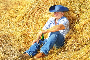 A man cowboy in a hat and jeans and a checkered rough shirt is resting after harvesting  drinking from a bottle of beer on a haystack