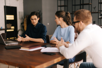 Young business man tells his colleagues about the project in modern office meeting against large panoramic window. Young man and woman listen to the speaker. Concept of office life.