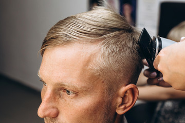 Fototapeta premium A blond hair man is hair cutting with barber in the barbershop. Haircut with an electric razor. Self-care. Close-up haircut. High quality photo