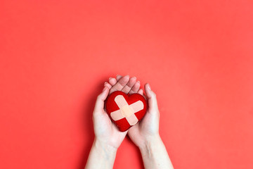 Heart with plaster in female hands on red background.