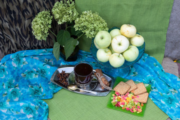 View from above. There is a glass of tea in a cup holder on a metal tray. Nearby is a plate of cookies and candied fruits, a vase of apples and a bouquet of hydrangea blossoms.