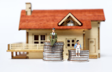 A miniature man and woman sitting on a pile of coins in front of a miniature wooden house.
