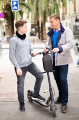 Fototapeta na wymiar Portrait of friendly adult man talking to his teenage son standing with electric scooter outdoors in autumn day..