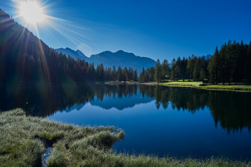 Obraz premium Sun on a crystal blue sky. Early morning in the Alps. Reflections of trees, mountains, sky in a mountain lake.