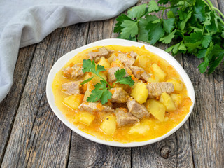 Homemade stew with meat and vegetables on a wooden background with grey linen towel and parsley. Tasty and nourishing lunch. Pork meat stewed with potatoes, carrots and spices, hungarian goulash