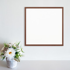 Wood frame mockup on white wall with pot of flowers on white surface. Dark wood. Copy space.