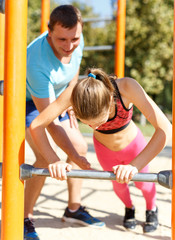 Fototapeta premium Sporty preteen girl doing push-ups on gymnastics bar during workout with her father on summer sports ground..
